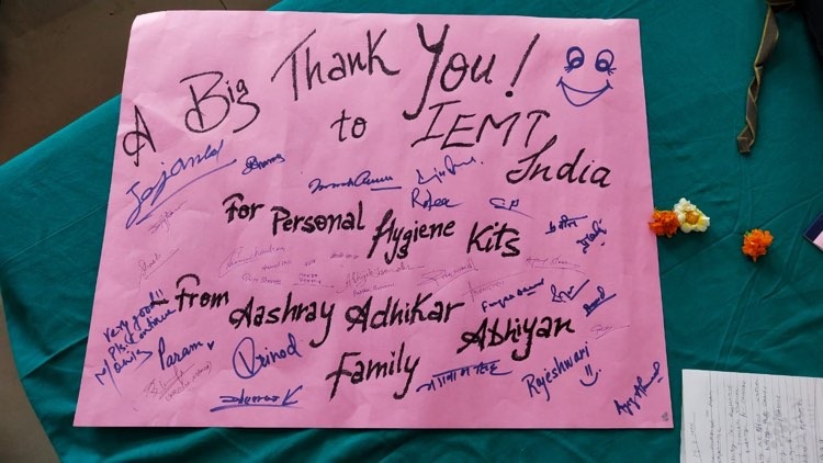 A pink poster on a teal table reads A Big Thank You! to IEMT, India for Personal Hygiene Kits from Akshay Adhikar Family...