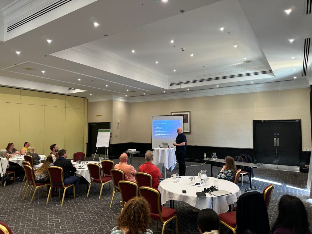 A man stands at the front of a conference room giving a presentation on trauma and EMDR to a seated audience. He speaks...