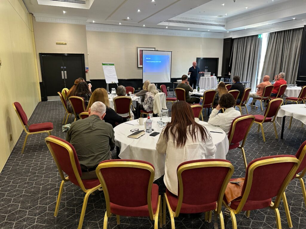 A group of people sit at round tables in a conference room, watching a presentation on trauma and PTSD projected on a...