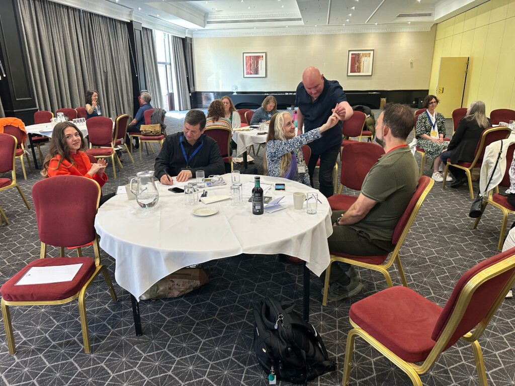 People are seated in small groups at round tables in a conference room, engaged in discussion about trauma and PTSD. One...