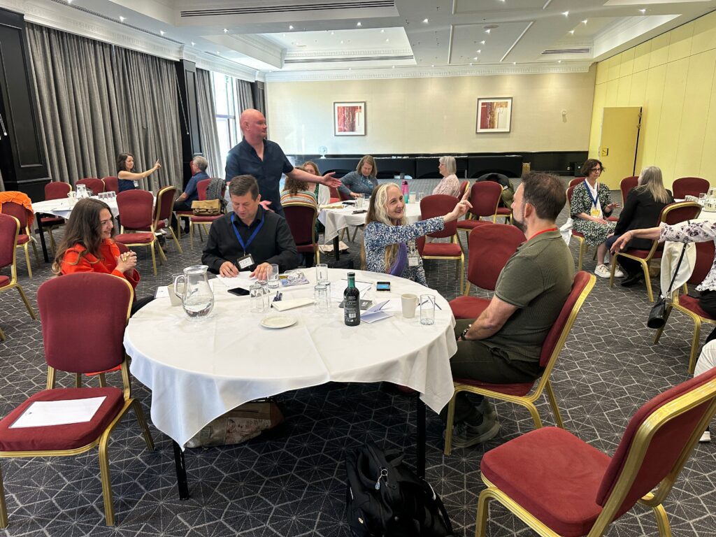People sit and talk in small groups around round tables in a conference room with red chairs and white tablecloths,...