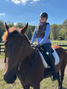 A person wearing a helmet and riding gear smiles whilst sitting on a brown horse outdoors, finding joy and calm—an uplifting moment that can support healing for those managing trauma or PTSD. Trees, a wooden fence, and clear blue sky fill the background.