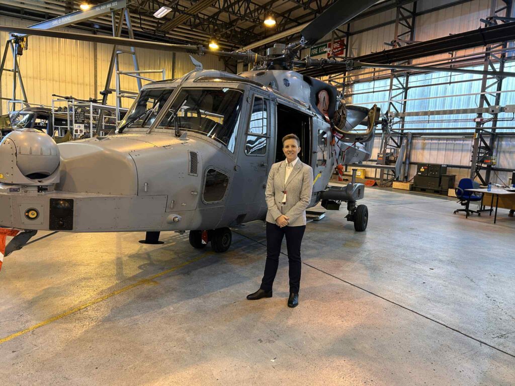 A person in business attire stands and smiles in front of a grey military helicopter inside a large, well lit hangar,...