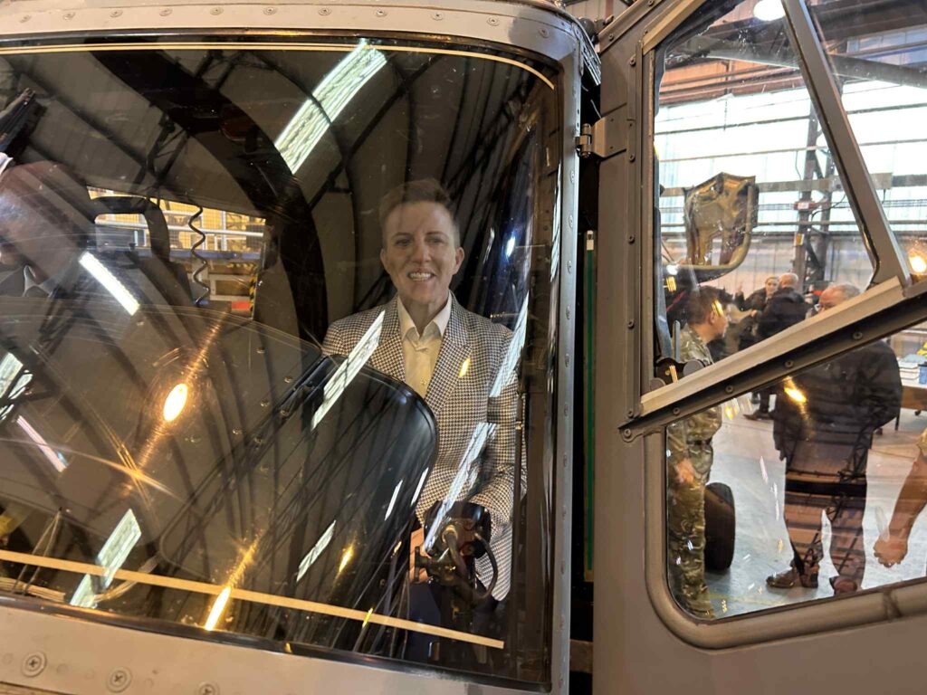 A person in a light coloured chequered jacket smiles while standing inside the cockpit of an aeroplane, visible through...