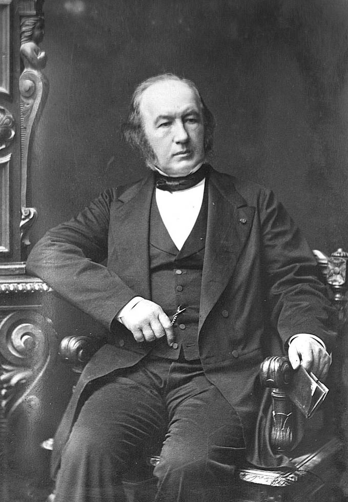 A seated man in formal 19th century attire, with sideburns, holding spectacles in one hand and a book or document on...