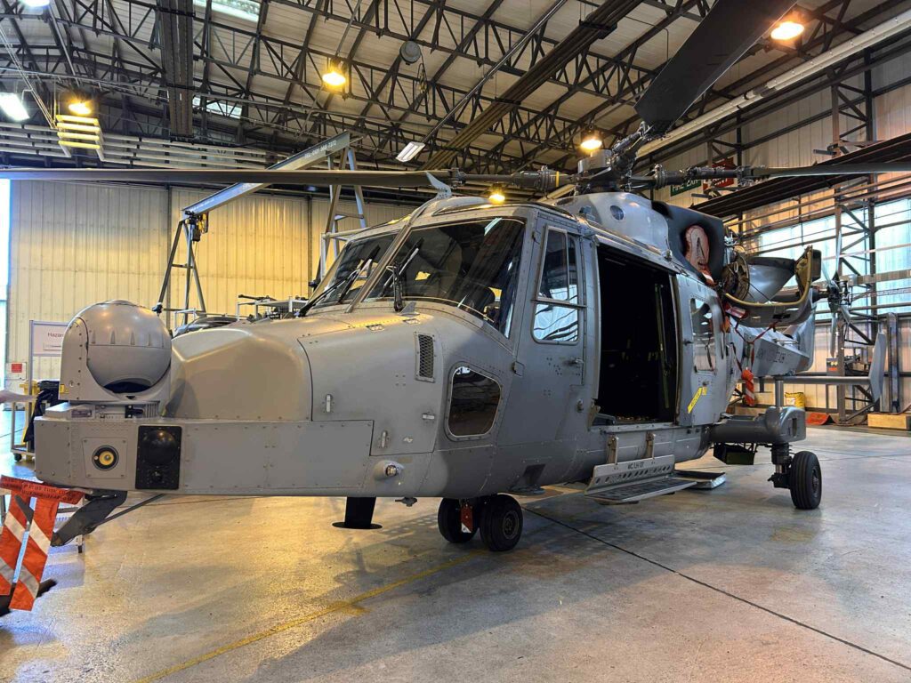 A grey military helicopter is parked inside a large hangar with its side door open, under bright industrial lighting and...