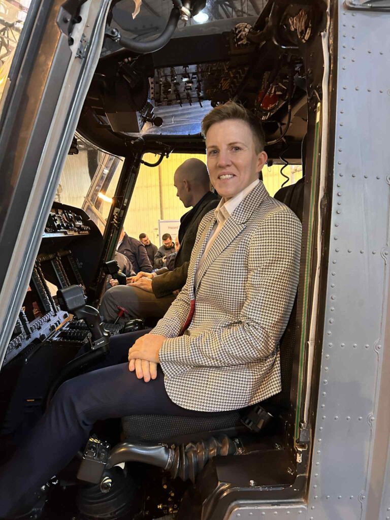 A person wearing a light chequered blazer and dark trousers sits in the cockpit of an aeroplane, smiling at the camera—a...