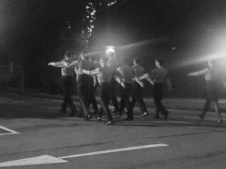 Black-and-white photograph of uniformed cadets marching outdoors during the evening event.